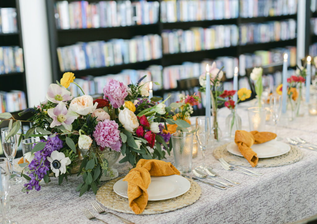 A table with plates and flowers sits in front of a wall of books. 