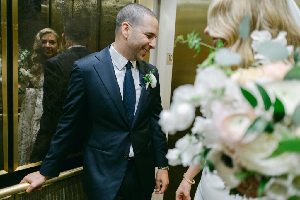 A bride is reflected in an elevator while the groom blushes.