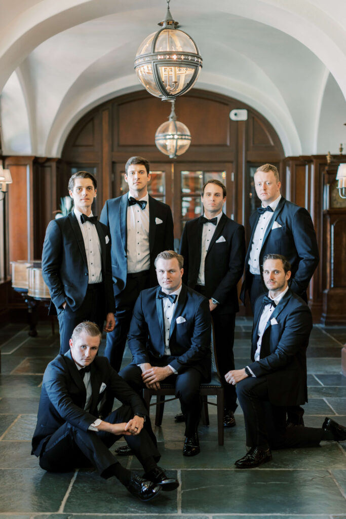 A groom sits in a chair while his best men stand around him. 
