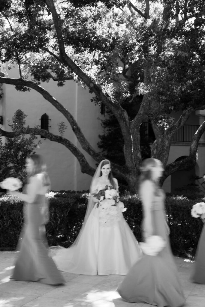 A bride stands still while blurs of bridesmaids walk around her. 