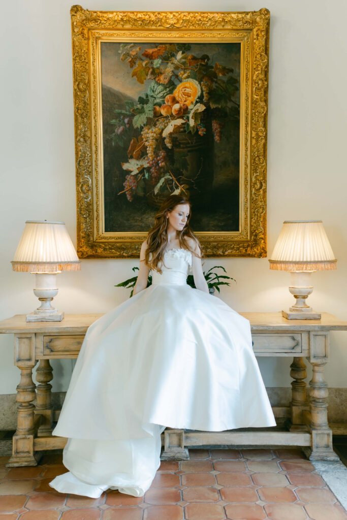 A bride sits on an antique desk, between two lamps and in front of a painting. 