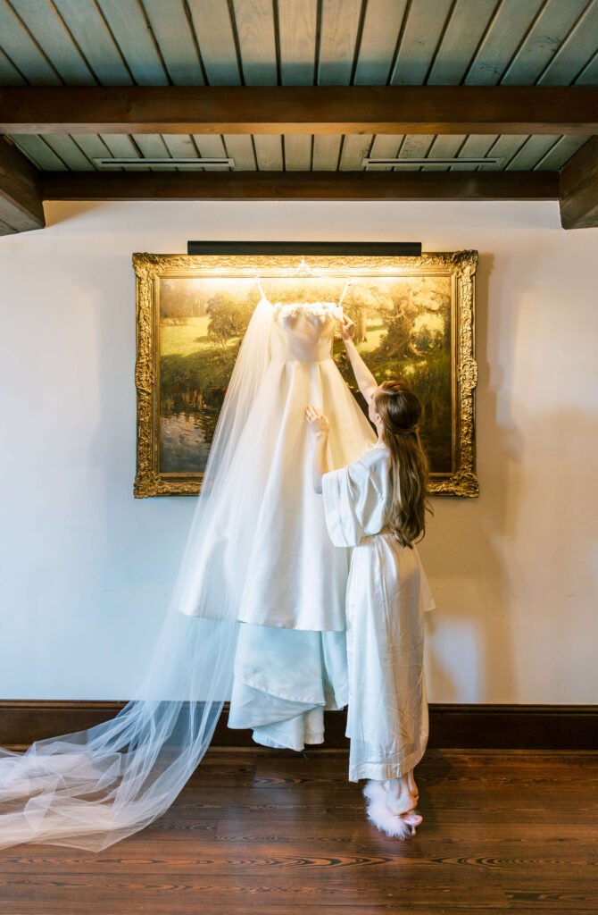 A bride admiring her wedding dress as it hangs over a painting. 